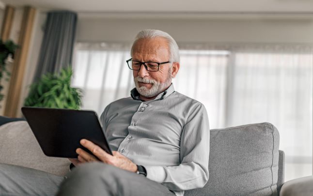 Äldre man sitter i soffan och ler med en ipad i handen